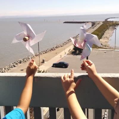 Children's hands hold up pinwheels, with bay jetty in the background.